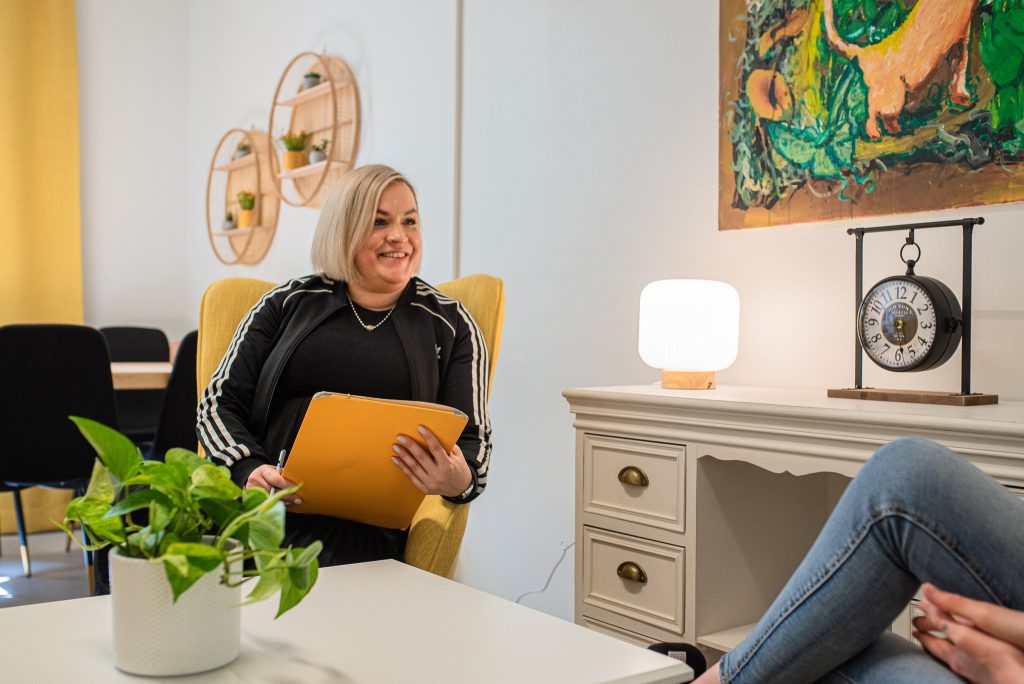 Sosionomi Henna Raiski keskustelee nuoren asiakkaan kanssa hymyillen Aarrerannan vastaanottohuoneessa keltainen lehtiö kädessään.
